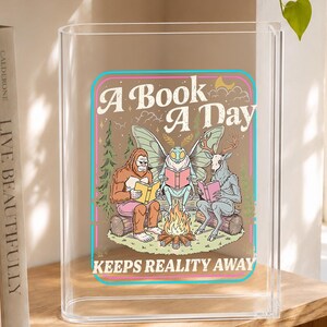 Può includere: Un leggio in acrilico trasparente con una grafica colorata che raffigura un sasquatch, una falena e un cervo seduti attorno a un falò a leggere. Il testo A BOOK A DAY KEEPS REALITY AWAY è scritto in un carattere retrò con pini e una luna.