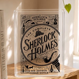 May include: A clear acrylic book display stand featuring a black graphic print of The Complete Sherlock Holmes Collection. The design includes illustrations of a deerstalker hat, a pipe, a violin, and Big Ben, with text by Sir Arthur Conan Doyle.