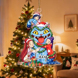 May include: A Christmas ornament featuring a blue cartoon cat with yellow eyes, a red tongue, and a Santa hat. Three figures are hugging the cat, and the name "Lucy" is written on the cat. The ornament hangs in front of a decorated Christmas tree.
