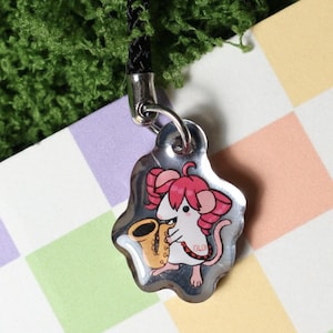 May include: A silver-colored charm with a cartoon mouse playing a saxophone. The mouse has pink ears and a pink tail. The charm is attached to a black cord. The background is a colorful checkered pattern.