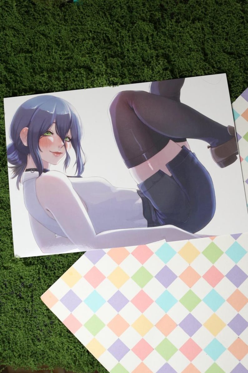 May include: A print featuring an anime-style illustration of a person with blue hair and green eyes, wearing a white top, dark shorts, and black tights. The person is lying down, with a playful expression. The print is on a white background, with a colorful patterned border.