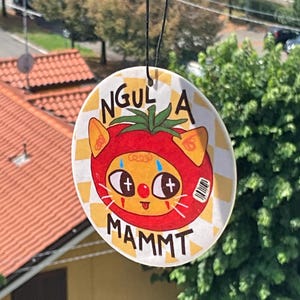 May include: A round, hanging ornament featuring a cartoon cat dressed as a tomato. The cat has orange ears and a red tomato costume. The ornament has the words "NGUL A MAMMT" in black text, set against a yellow and white checkered background.