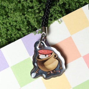 May include: A clear, glossy charm featuring a cartoon mouse in a cooking pot with a red lid. The charm is attached to a black braided cord with a silver-colored metal clasp. The background is a colorful checkered pattern.