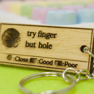 May include: A rectangular wooden keychain with the text "try finger but hole". The keychain has a fingerprint graphic and a rating scale from Close to Poor. A silver keyring is attached.