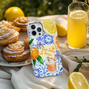 May include: A phone case featuring a citrus fruit design with the text "Que Sera Sera". The case has a white and blue pattern. It is next to a glass of orange juice, pastries, and lemons.