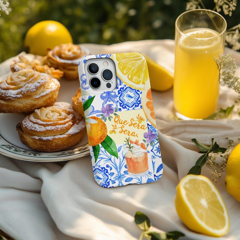 May include: A phone case featuring a citrus fruit design with the text "Que Sera Sera". The case has a white and blue pattern. It is next to a glass of orange juice, pastries, and lemons.