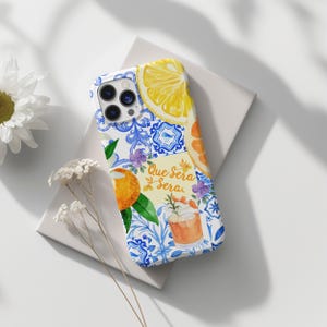 May include: A white phone case with a vibrant design of lemons, oranges, and blue and white tile patterns. The case features the text "Que Sera Sera" and a cocktail illustration.