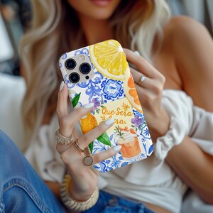May include: A white phone case with a vibrant design of lemons, oranges, and blue floral motifs. The case displays the text "Que Sera Sera". The phone case has a clear camera lens opening.