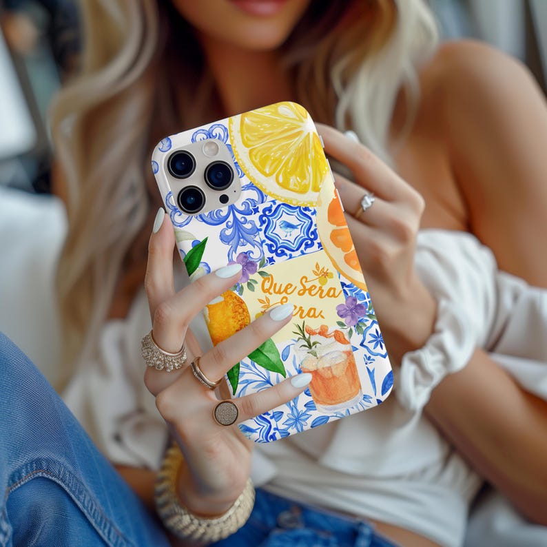 May include: A white phone case with a vibrant design of lemons, oranges, and blue floral motifs. The case displays the text "Que Sera Sera". The phone case has a clear camera lens opening.