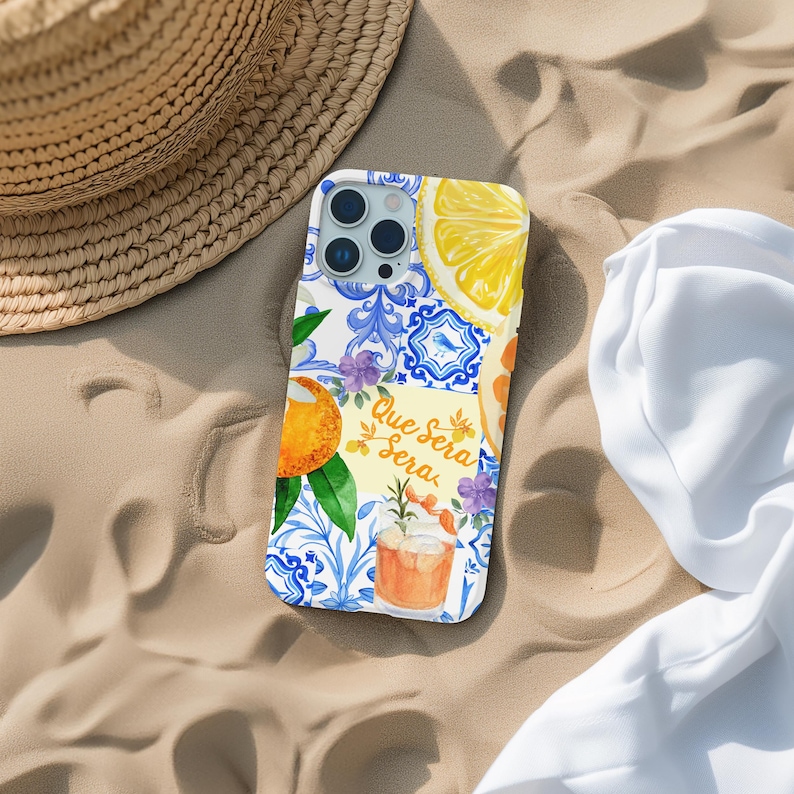 May include: A white phone case with a vibrant design including a lemon slice, orange, blue floral tiles, and the text "Que Sera Sera." The case rests on sand beside a straw hat and white fabric.