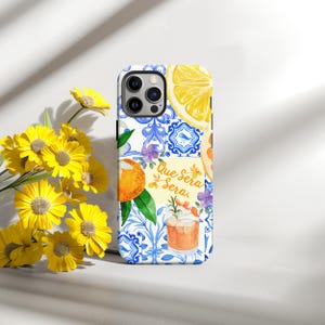 May include: A phone case with a vibrant design featuring a lemon slice, orange slice, cocktail, and blue and white tile pattern. The case has the text "Que Sera Sera" and is surrounded by yellow flowers.