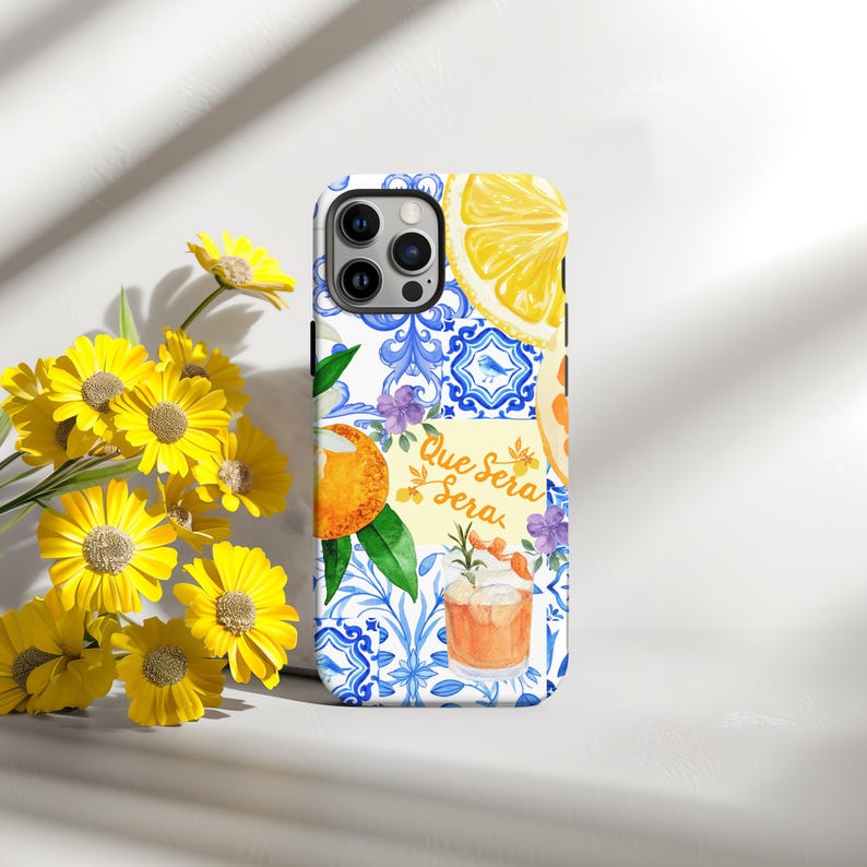 May include: A phone case with a vibrant design featuring a lemon slice, orange slice, cocktail, and blue and white tile pattern. The case has the text "Que Sera Sera" and is surrounded by yellow flowers.