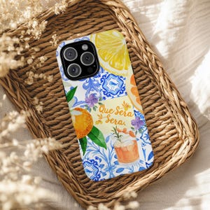 May include: A white phone case with a colourful design featuring lemon and orange slices, blue and white floral patterns, and the text "Que Sera Sera". The case is on a woven basket.