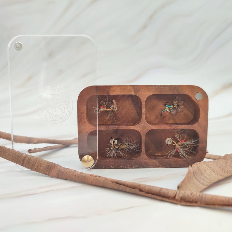 Puede incluir: Una caja de pesca con mosca de madera con cuatro compartimentos, cada uno con una mosca diferente. La caja est&aacute; hecha de madera oscura y tiene una tapa de acr&iacute;lico transparente.