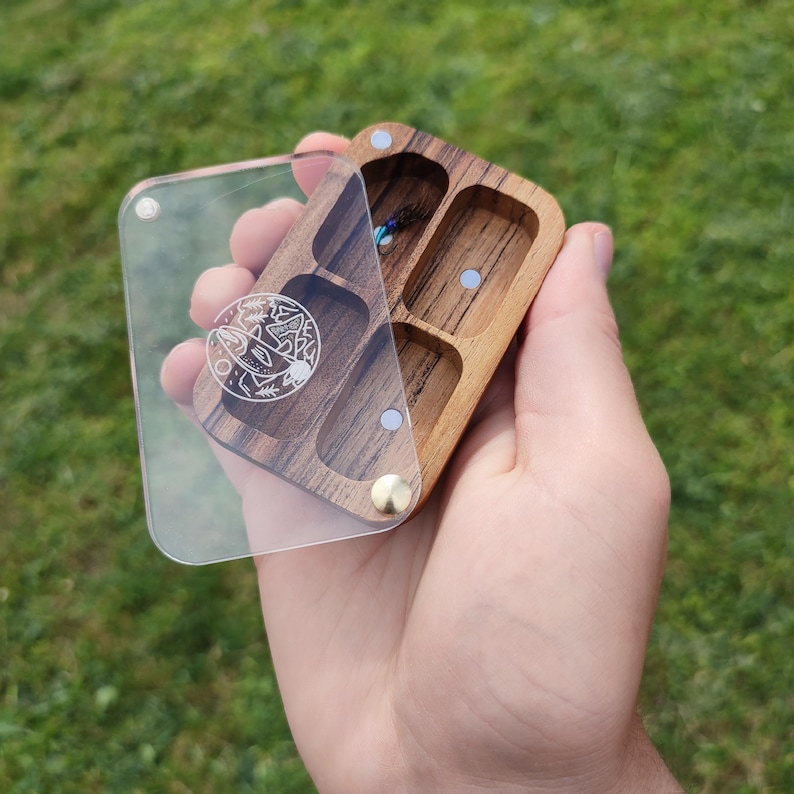 Puede incluir: Una caja de madera con una tapa de acr&iacute;lico transparente. La caja tiene cuatro compartimentos y est&aacute; grabada con un dise&ntilde;o de un pez saltando fuera del agua. La caja es perfecta para guardar objetos peque&ntilde;os como joyas o se&ntilde;uelos de pesca.