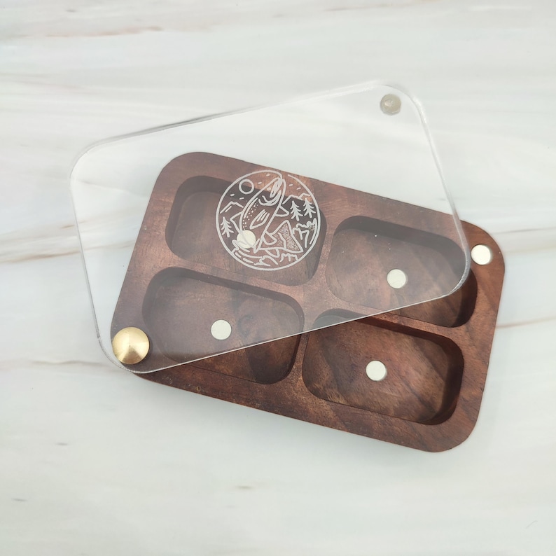 Puede incluir: Una caja de madera con una tapa de acr&iacute;lico transparente. La caja tiene cinco compartimentos y un dise&ntilde;o de un pez saltando fuera del agua con monta&ntilde;as en el fondo. La tapa tiene una bisagra de lat&oacute;n.