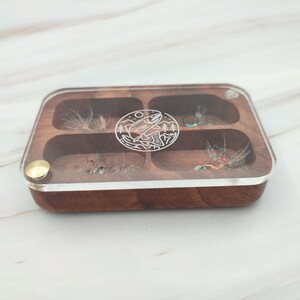 Puede incluir: Una caja de pesca con mosca de madera con una tapa de acr&iacute;lico transparente. La caja tiene seis compartimentos y presenta un dise&ntilde;o grabado de un pez y &aacute;rboles.