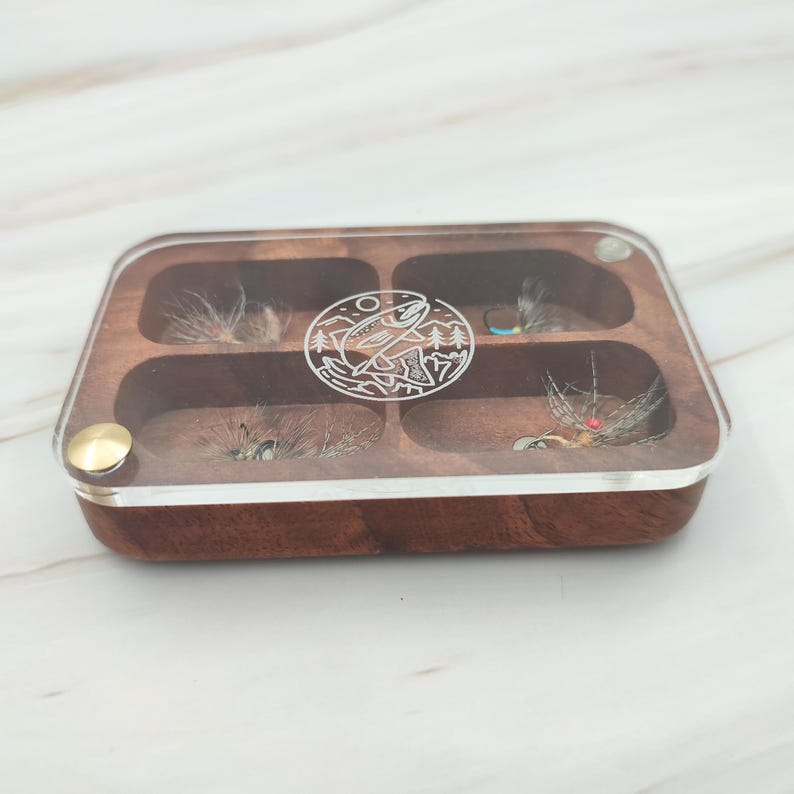 Puede incluir: Una caja de pesca con mosca de madera con una tapa de acr&iacute;lico transparente. La caja tiene seis compartimentos y presenta un dise&ntilde;o grabado de un pez y &aacute;rboles.