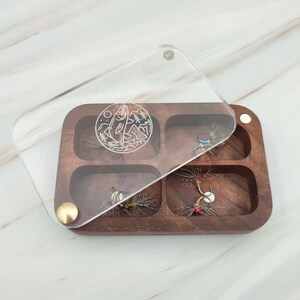 Puede incluir: Una caja de pesca con mosca de madera con seis compartimentos para guardar moscas. La caja tiene una tapa de acr&iacute;lico transparente con un dise&ntilde;o grabado con l&aacute;ser de un pez saltando fuera del agua.