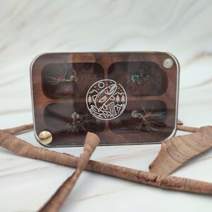 Puede incluir: Una caja de pesca con mosca de madera con seis compartimentos para guardar moscas. La caja tiene una tapa de acr&iacute;lico transparente y un cierre de lat&oacute;n. La caja est&aacute; decorada con un dise&ntilde;o grabado con l&aacute;ser de un pez saltando fuera del agua.