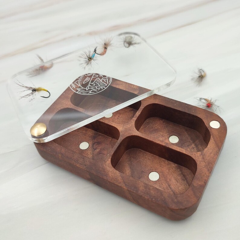 Puede incluir: Una caja de pesca con mosca de madera con una tapa de acr&iacute;lico transparente. La caja tiene cuatro compartimentos y est&aacute; forrada con imanes. La tapa tiene un dise&ntilde;o decorativo de un pez y una mosca.