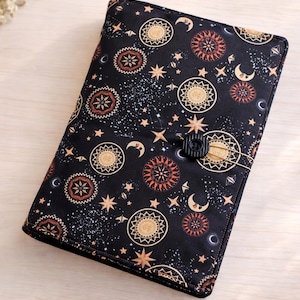 May include: Black book cover with a celestial pattern featuring suns, moons, and stars in gold, orange, and cream. The cover has a button closure and is made of fabric. The text "@Bookshelf.at" is visible at the bottom.