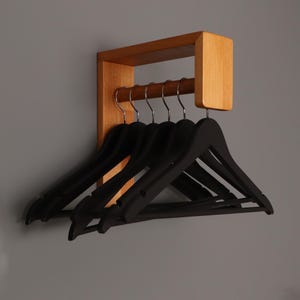 Könnte beinhalten: Ein Wandhalter aus Holz mit natürlicher Oberfläche. Der Halter hat eine horizontale Stange und ein Regal. Sechs schwarze Plastikbügel hängen an der Stange.