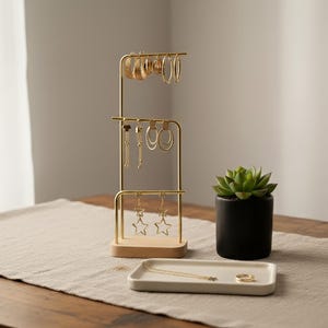 Può includere: Un espositore per gioielli in metallo color oro a tre livelli, che mostra vari orecchini. Sono presenti anche un piccolo vaso nero con una succulenta e un vassoio bianco con gioielli. L'espositore è su una base di legno.