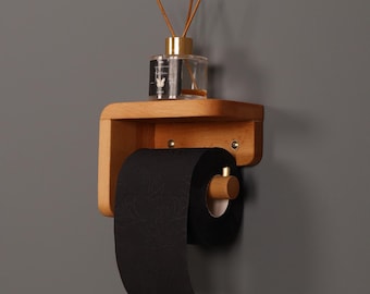May include: A wall-mounted light wood toilet paper holder with a flat top shelf. A roll of black toilet paper with a subtle pattern is mounted on the wooden dowel. A clear glass reed diffuser bottle with wooden sticks sits on the top shelf.