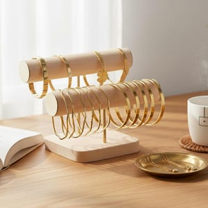 May include: A wooden jewellery display stand with two levels, holding gold bracelets and bangles. The stand has a natural wood finish with gold accents. A small gold dish with necklaces sits on the table. A cup and open book are also visible.