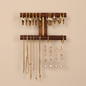 Könnte beinhalten: Ein hölzerner Schmuck-Organizer mit goldfarbenen Armbändern, Halsketten und Ohrringen. Der Organizer hat zwei horizontale Stangen für Armbänder und einen vertikalen Abschnitt für Halsketten und Ohrringe. Der Schmuck wird vor einem neutralen Hintergrund präsentiert.
