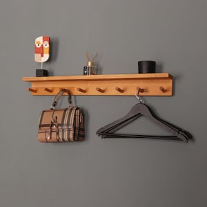 Könnte beinhalten: Ein Wand-Kleiderständer aus Holz mit sechs Haken. Eine braune und beige karierte Handtasche hängt an einem Haken. Drei schwarze Kleiderbügel hängen an einem anderen Haken.
