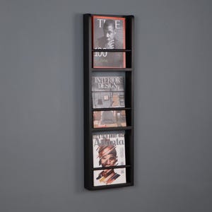 May include: A tall, rectangular black wall-mounted magazine rack featuring four horizontal shelves. The rack holds four magazines, including issues titled Time, Interior Design, and Atlanta, which are displayed vertically in the open frame.