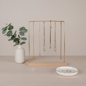 May include: A gold-coloured jewellery display stand with several gold necklaces hanging from it. A small white vase with green leaves is to the left. A small white dish with gold trim and two rings sits in front of the stand.