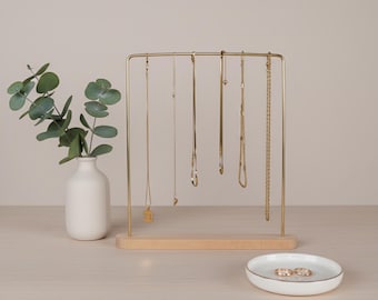 Puede incluir: Un expositor de joyas de color dorado con varios collares dorados colgando. A la izquierda, un pequeño jarrón blanco con hojas verdes. Delante del expositor hay un plato blanco pequeño con ribete dorado y dos anillos.