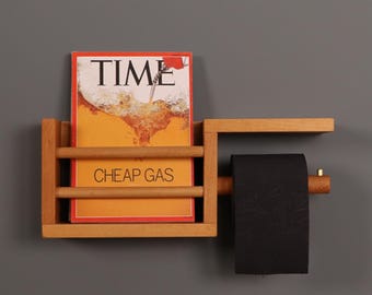 May include: A light brown wooden wall-mounted organizer featuring a magazine rack holding a TIME magazine with the text CHEAP GAS and a horizontal bar supporting a roll of black toilet paper with a small brass-colored end cap detail.