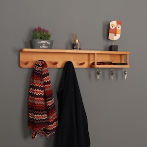 May include: A wooden wall shelf with pegs, holding a potted plant, a decorative face sculpture, and a small shelf for keys and glasses. A patterned scarf and a black coat hang from the pegs. The text "FLOWERS & GARDEN" is visible. The shelf is made of wood.