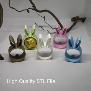 以下が含まれることがあります： 5つの異なる色の3Dプリントされたウサギの形をしたホルダー。茶色、緑、白、ピンク、青。ホルダーは、キャンディーや卵などの小さなアイテムを保持するように設計されています。ホルダーの下には「High Quality STL File」というテキストが印刷されています。