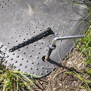 May include: A black plastic ground stake with a spiral design and a metal hex key are shown on a textured black surface. The stake is designed to be screwed into the ground. The hex key is inserted into a metal bolt.