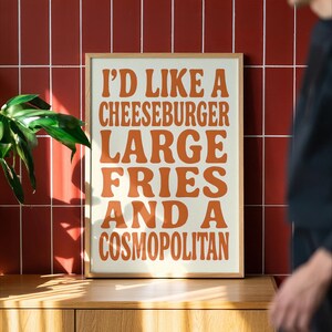 Framed poster with bold retro orange typography reading “I’D LIKE A CHEESEBURGER, LARGE FRIES AND A COSMOPOLITAN,” set on a cream background and displayed against a red tiled wall with warm sunlight and soft shadows, created by Nelly’s Digital Studio.