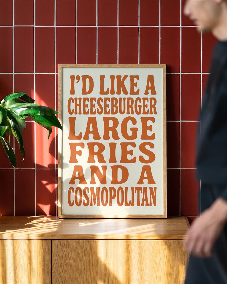 Framed poster with bold retro orange typography reading “I’D LIKE A CHEESEBURGER, LARGE FRIES AND A COSMOPOLITAN,” set on a cream background and displayed against a red tiled wall with warm sunlight and soft shadows, created by Nelly’s Digital Studio.