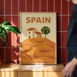 May include: A framed art print with the word "SPAIN" in orange letters. The artwork depicts a landscape with two houses, a tree, and rolling hills in shades of orange and brown. The frame is made of wood.