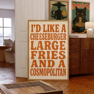 Retro kitchen wall art featuring bold orange typography that reads “I’d like a cheeseburger, large fries, and a cosmopolitan,” styled in a cozy mid-century space with wood and eclectic decor