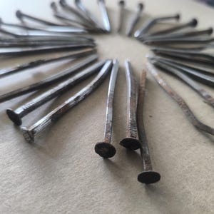 Può includere: Primo piano di una collezione di chiodi metallici disposti a cerchio. I chiodi hanno un aspetto scuro e invecchiato con ruggine e usura visibili. Le teste dei chiodi sono piatte e circolari.