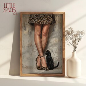 May include: A framed art print depicting a pair of legs wearing a leopard print skirt and matching leopard print high heels with ankle straps. A small black cat sits beside the legs against a textured, neutral-toned wall background.