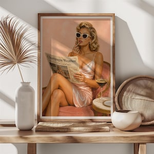 May include: A framed wall art print featuring a person with blonde hair wearing white sunglasses and a light pink slip dress, sitting in a chair while reading a newspaper titled Champagne Weather and holding a martini glass with a dark liquid.