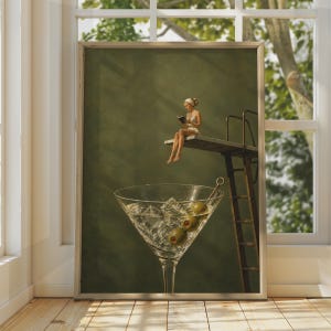 Op de afbeelding: Een ingelijste kunstdruk met een persoon in een wit badpak die op een duikplank zit en een boek leest boven een groot martiniglas gevuld met ijsblokjes en drie groene olijven aan een cocktailprikker, tegen een donker olijfgroene achtergrond.