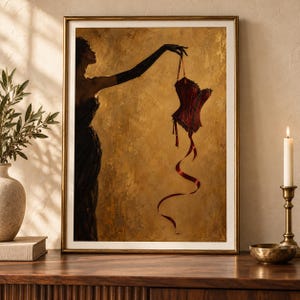 May include: A framed wall art print featuring a silhouette of a figure in a dark gown holding a deep red corset with long, flowing ribbons. The background is a textured, metallic gold color, and the entire composition is encased in a gold frame.