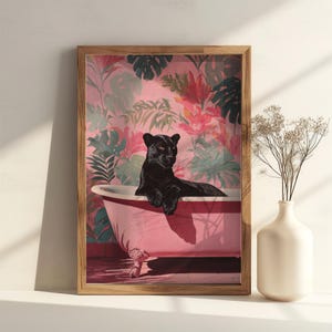 May include: A framed art print featuring a black panther lounging in a pink bathtub. The artwork has a tropical floral wallpaper background. The frame is made of light brown wood. A white vase with dried flowers is on the right.