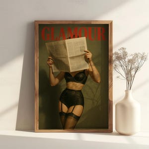 May include: A framed print showing a woman in black lingerie with a newspaper obscuring her face. The word "GLAMOUR" is printed in red at the top. The print is in a wooden frame and is displayed on a white surface.